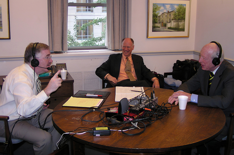 Image of three individuals in professional attire seated around a table with podcast equipment