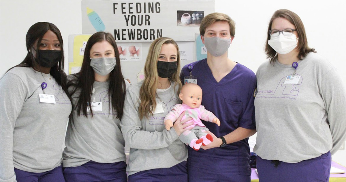 Nursing students stand in front of a posterboard explaining how to feed your newborn. They are all wearing masks, purple scrubs, and grey shirts.