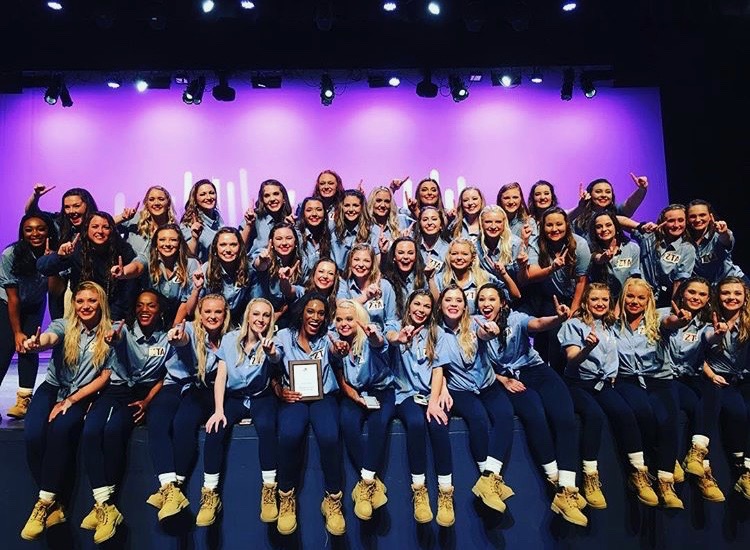 Large group of Zeta Tau Alpha members in denim shirts, jeans, and work boots pose on stage after performing in the University's step sing competition.