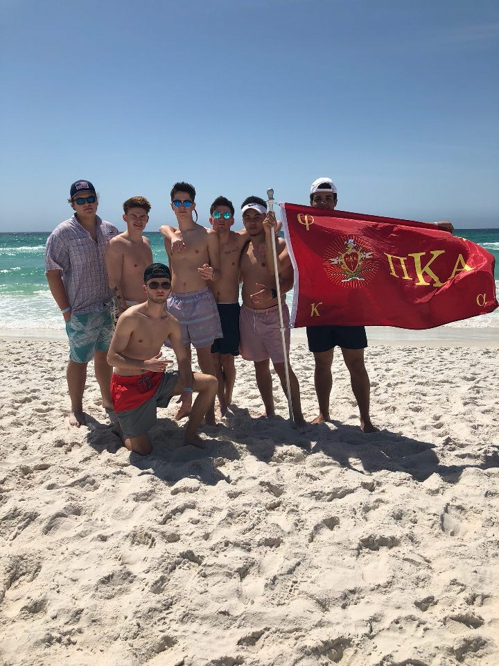 Men in swim trunks on a beach holding a red flag that says Pi Kappa Alpha in greek letters. 