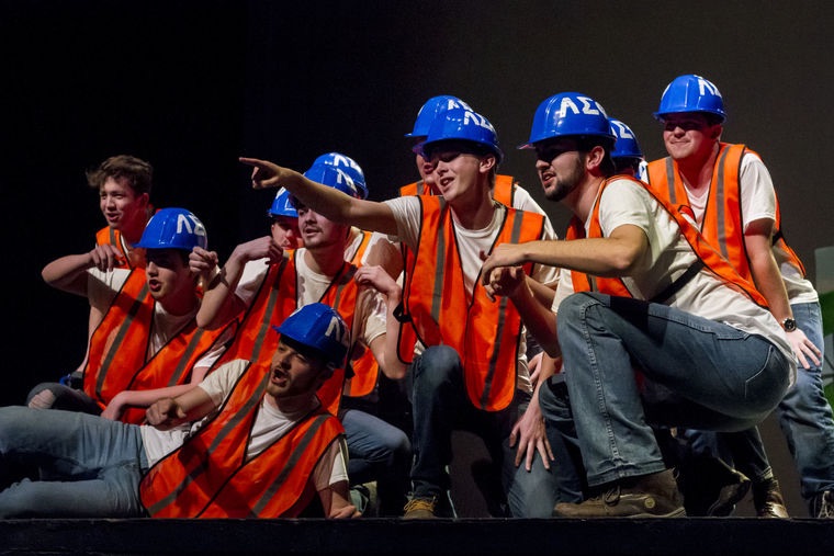Men in orange vests and blue hats gathered on stage during step sing performance, themed