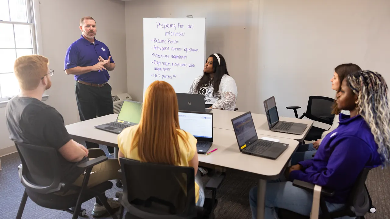 A faculty member leading a First-Year Experience class, students seated around a table with laptops and a whiteboard of notes behind him