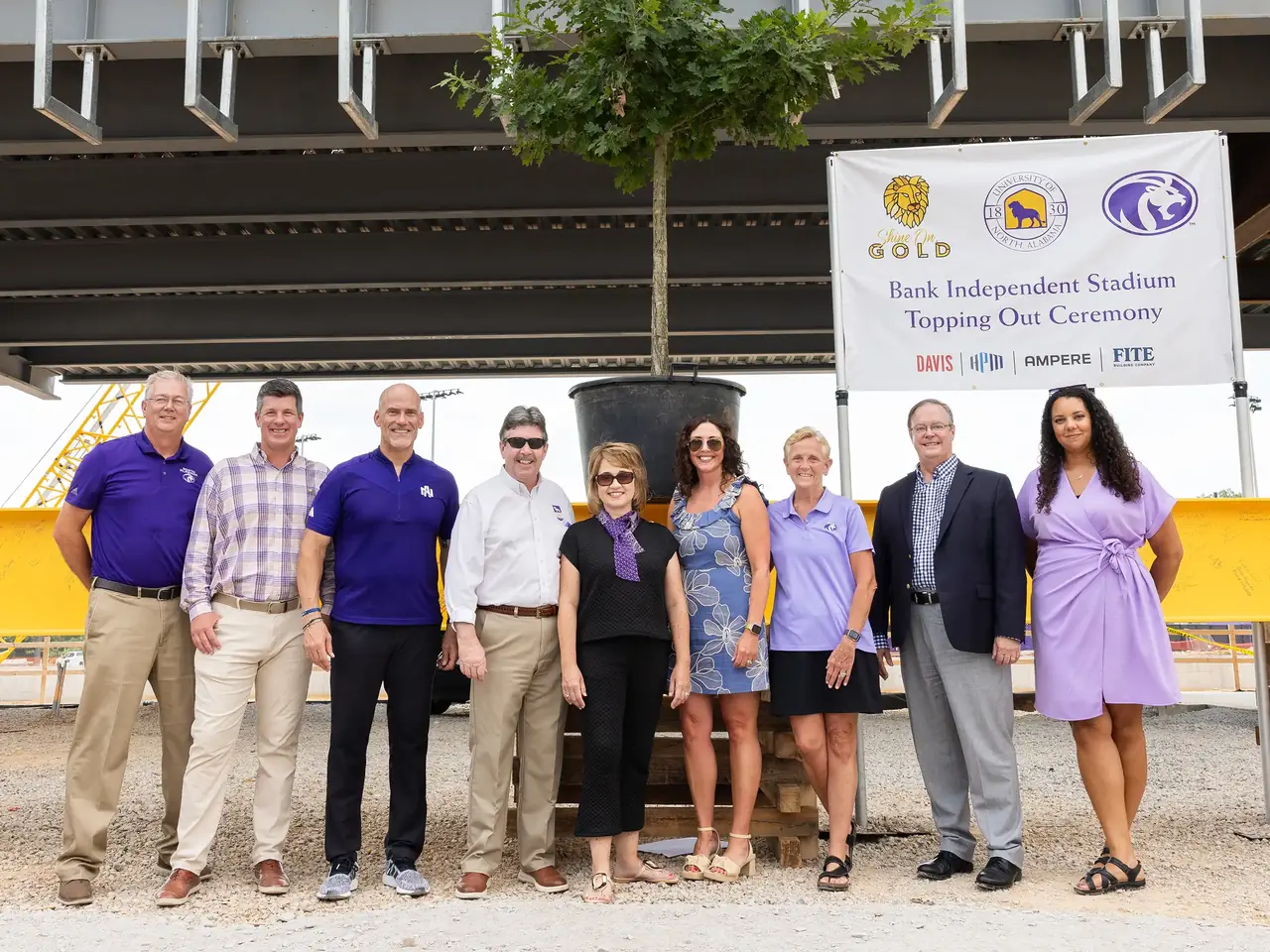 University of North Alabama leaders and the project's building partners at the Bank Independent Stadium topping-out ceremony, standing under the completed steel frame beside a ceremonial oak and a Shine On, Gold banner