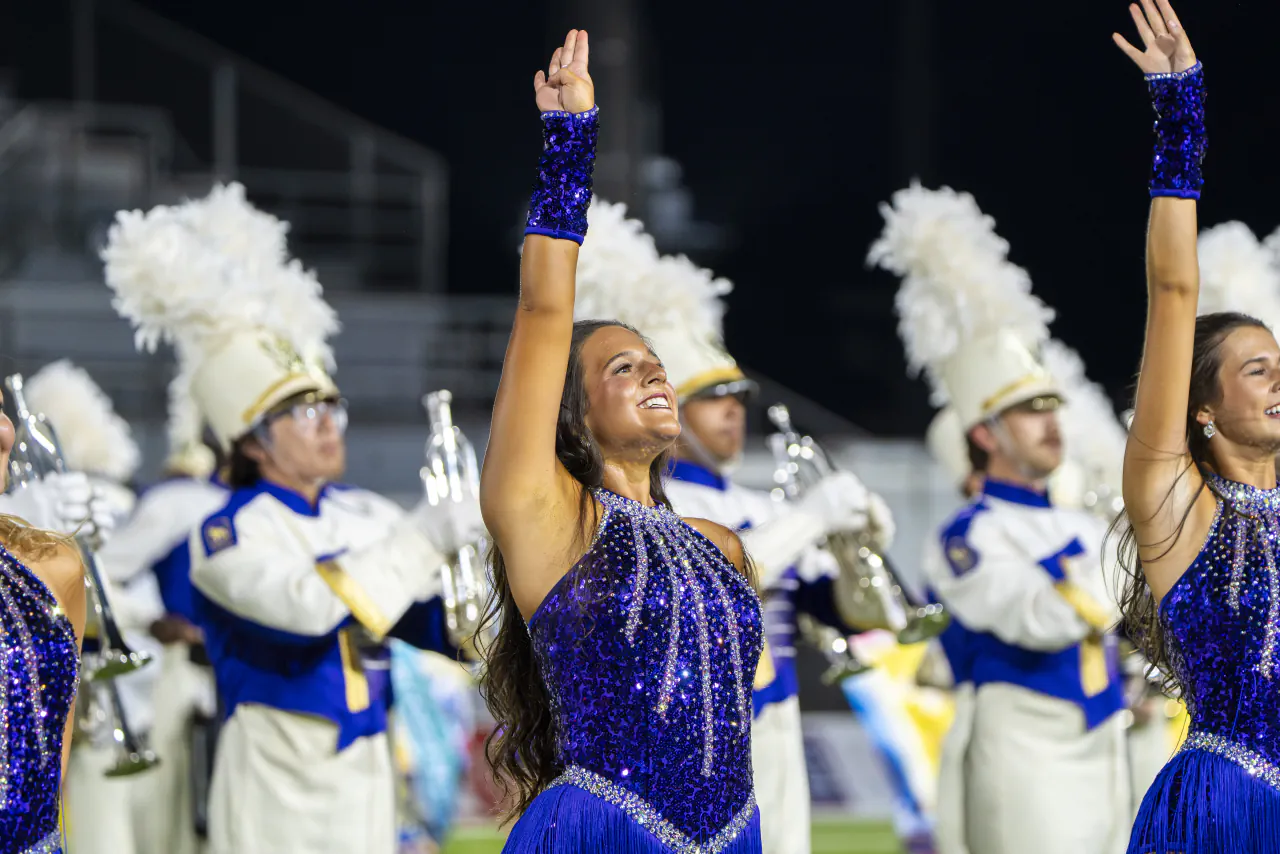 Golden Girls dancers with their arms raised in front of the marching band at a night football game