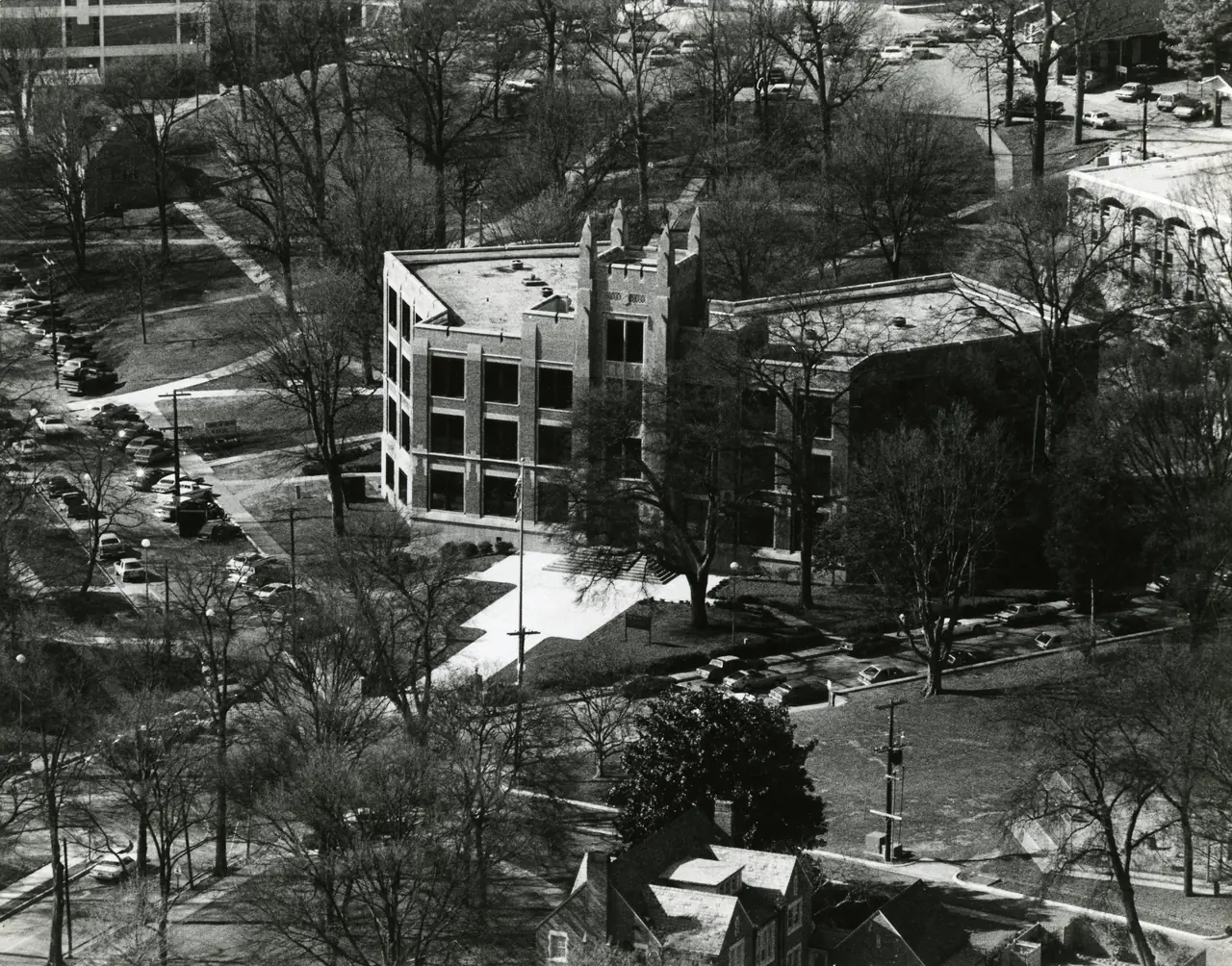 Aerial view of campus, a brick building at the center among trees, walkways and parking