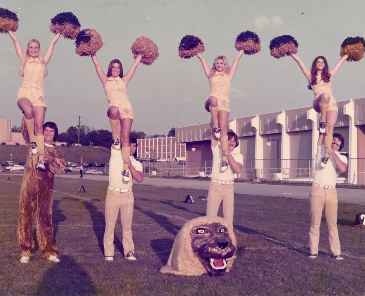 Cheerleaders in gold uniforms balanced on their partners&rsquo; shoulders, beside a costumed Leo and a giant lion head on the field