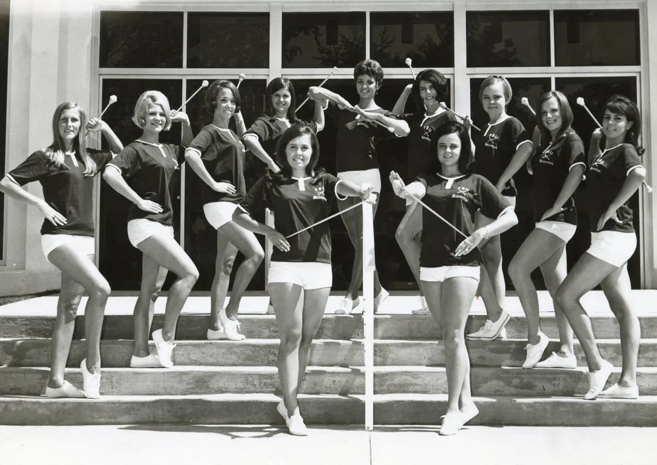 Eleven majorettes posed with batons on the steps of a campus building