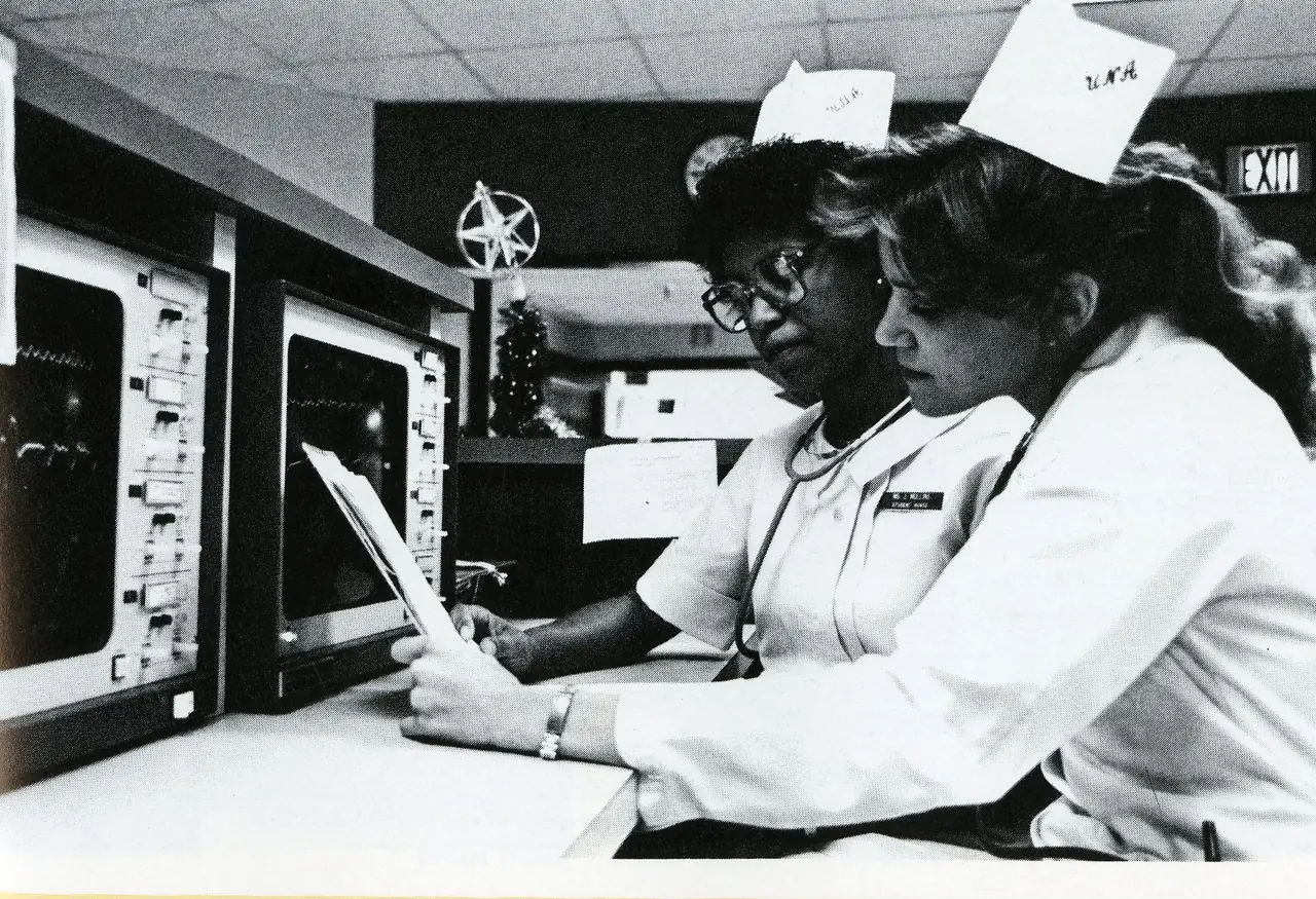 Two nursing students in white caps reviewing a chart beside monitoring equipment