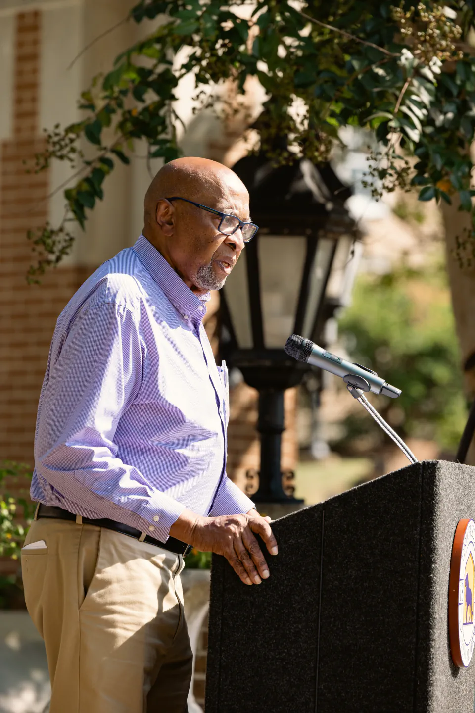 Wendell Gunn speaking from a lectern outdoors, a lamp post and greenery behind him