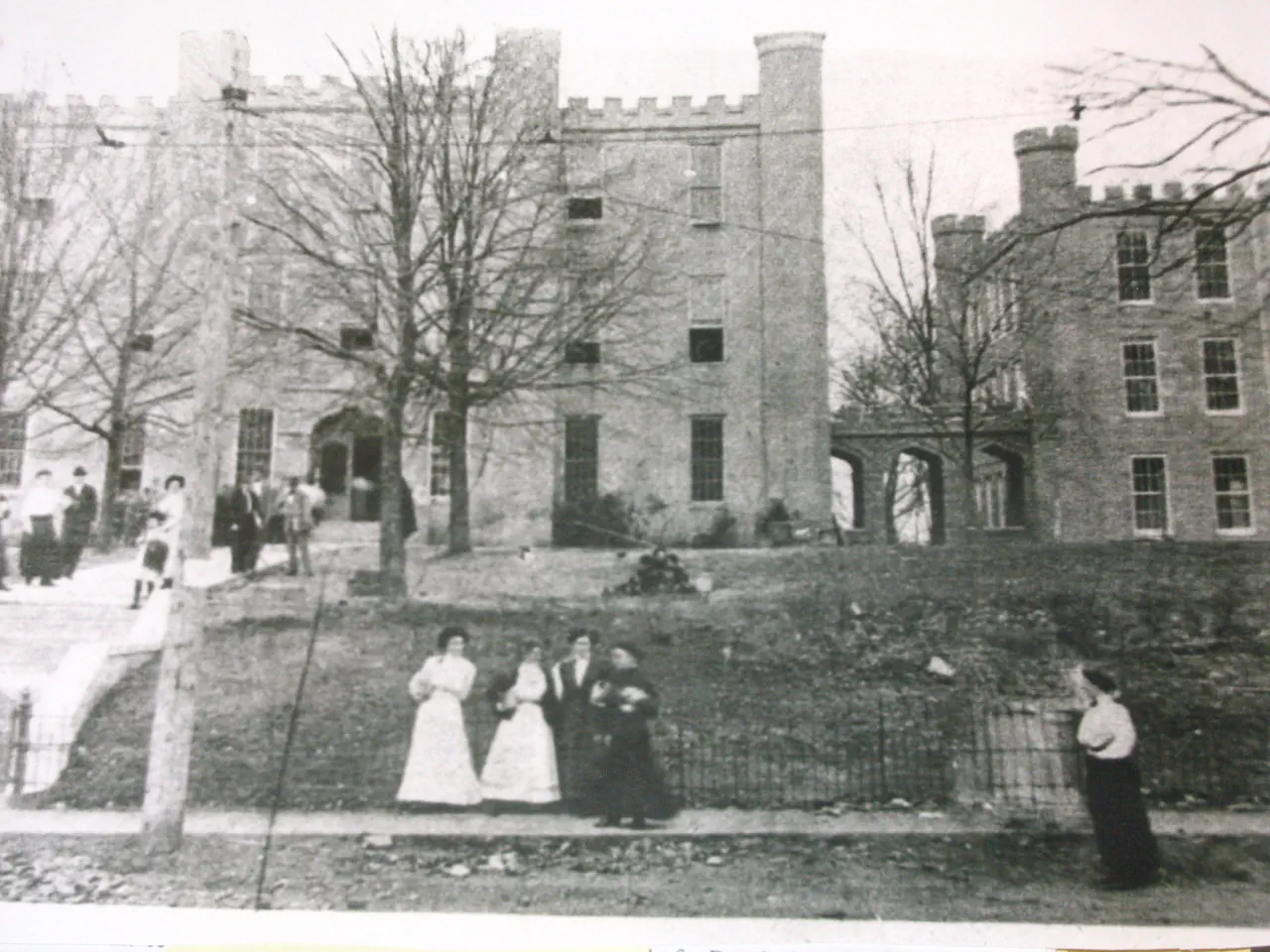 A group of women students in long white and dark dresses standing on the bank below Wesleyan Hall, with more students gathered at the entrance above